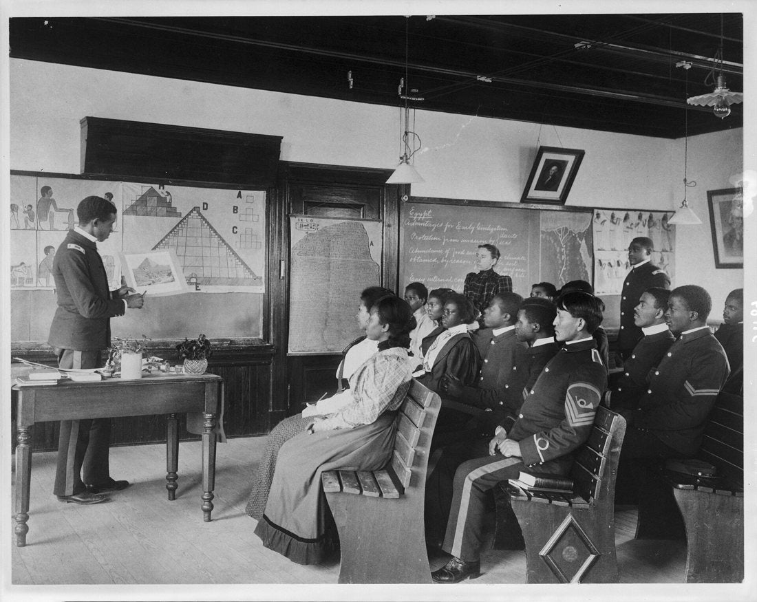Black-and-white photograph of students in uniform and civilian dress in a classroom at Hampton Institute, Virginia, in 1899, a man at a diagram of a pyramid